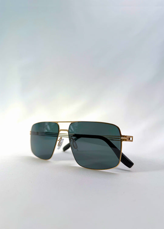 Side view of Axiom sunglasses in Antique Brass, featuring antique brass rectangular metal frame and green-grey polarized lenses, showing the lens shape and temple profile. 