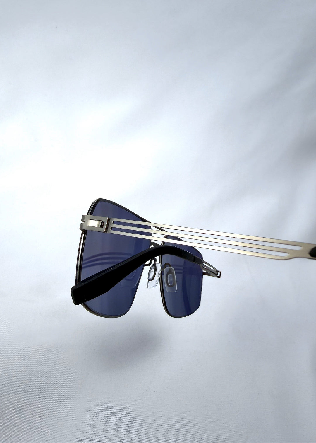 Close-up side detail view of Axiom sunglasses in Blue Graphite, featuring blue-graphite rectangular metal frame and blue tinted polarized lenses, showing the lens edge, hinge area, and temple detail. 