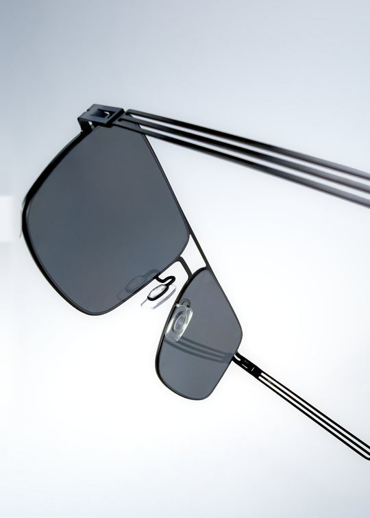 Close-up side detail view of Axiom sunglasses in Matte Black, featuring matte black rectangular metal frame and grey polarized lenses, showing the lens edge, hinge area, and temple detail. 