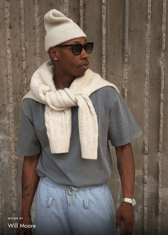 Will Moore wearing Cavalin sunglasses in Graphite Haze, with a cream knit beanie and sweater over a grey T-shirt against a textured concrete wall.