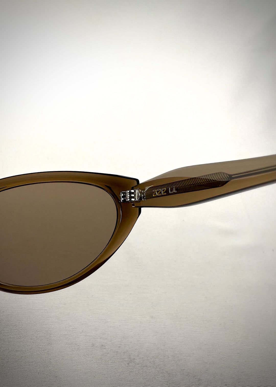 Close-up hinge detail view of Chara sunglasses in Champagne Brown, featuring a translucent champagne brown cat-eye acetate frame and UV400 lenses, showing the hinge construction and lens edge. 