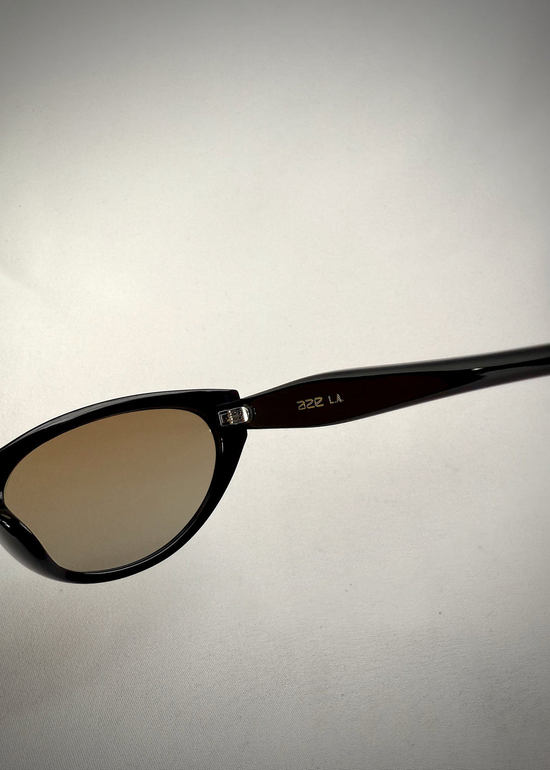 Close-up hinge detail view of Chara sunglasses in Gradient Amber, featuring a glossy black cat-eye acetate frame with amber gradient lenses, showing the hinge construction and lens edge. 