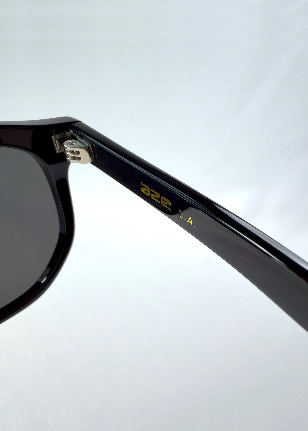 Close-up hinge detail view of Elara sunglasses in Obsidian Black, featuring black acetate frame and dark lenses, showing the hinge, lens edge, and temple detail. 