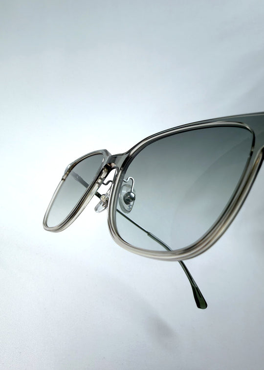 Close-up hinge detail view of Eon Drift sunglasses in Olive Shadow, featuring a translucent olive-gray frame and gray gradient lenses, showing the hinge construction, lens edge, and temple connection.