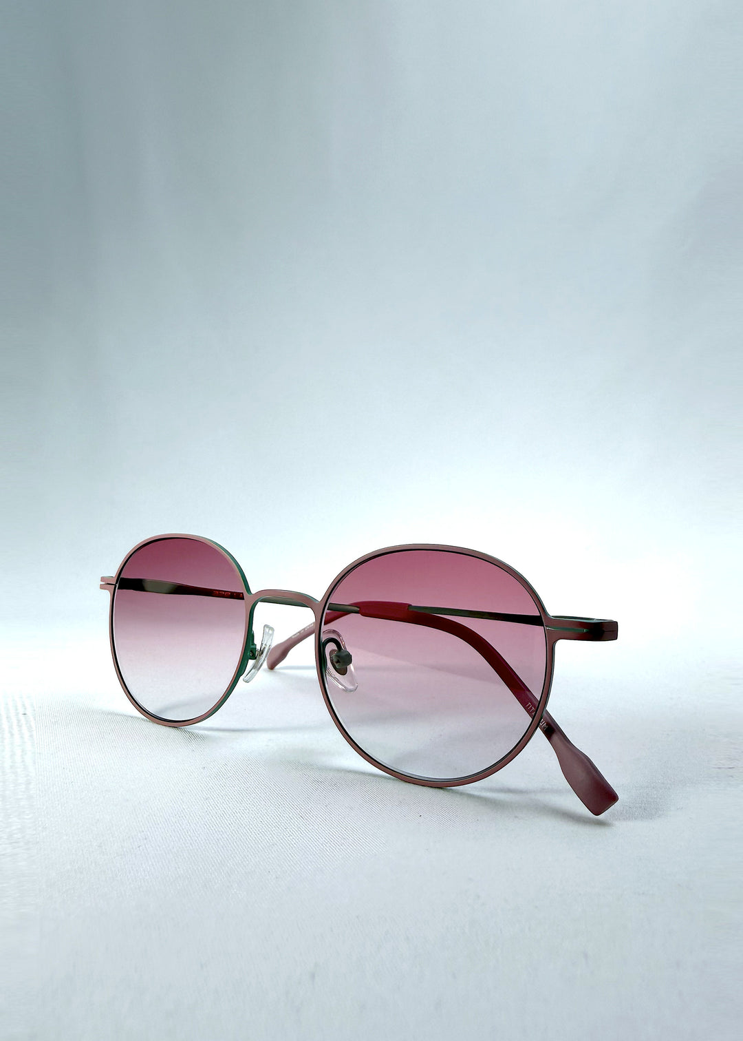 Folded view of AZE Liora Arc sunglasses in Mauve Halo, featuring a green-accented round frame and pink gradient lenses, showing the folded temples and frame profile.