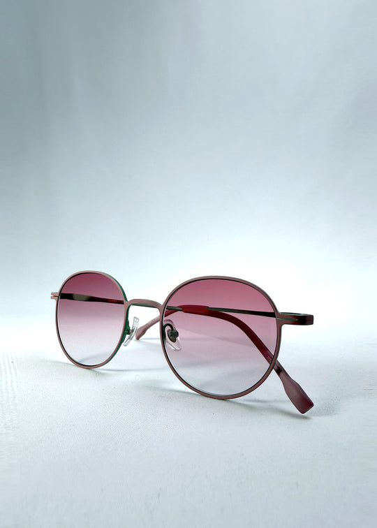 Folded view of AZE Liora Arc sunglasses in Mauve Halo, featuring a green-accented round frame and pink gradient lenses, showing the folded temples and frame profile.