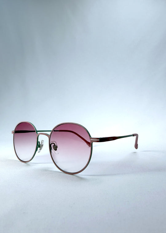 Three-quarter front view of AZE Liora Arc sunglasses in Mauve Halo, featuring a green-accented round frame and pink gradient lenses, showing the frame shape and temple profile.