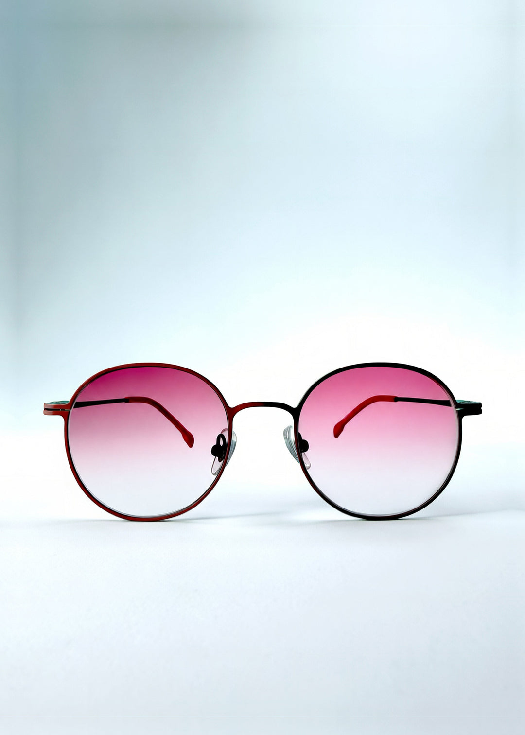 Front view of AZE Liora Arc sunglasses in Crimson Halo, featuring a deep crimson round metal frame with green temple accents and pink gradient lenses, showing the full front silhouette. 