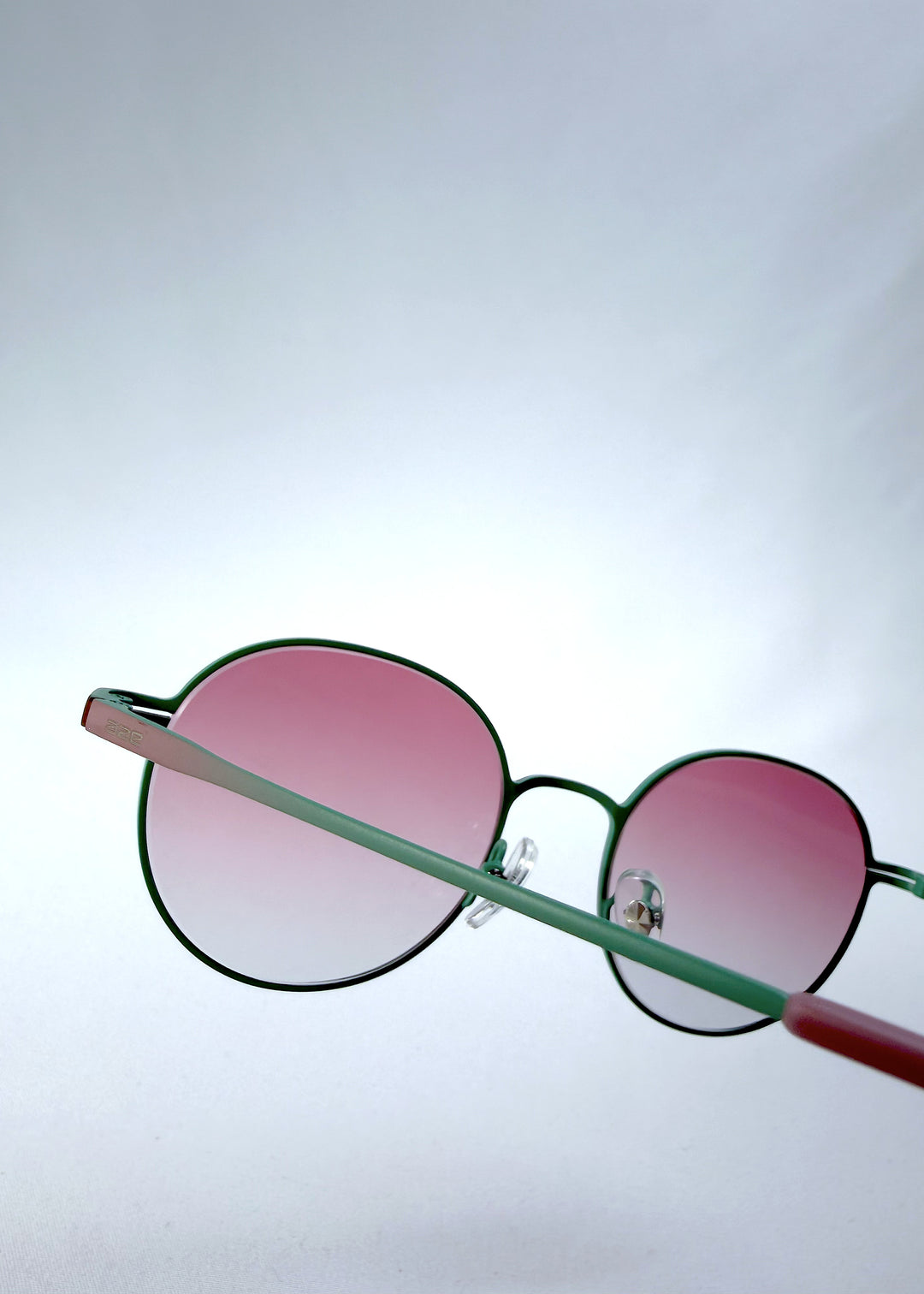 Close-up hinge detail view of AZE Liora Arc sunglasses in Mauve Halo, featuring a green-accented round frame and pink gradient lenses, showing the hinge construction, lens edge, and temple connection.