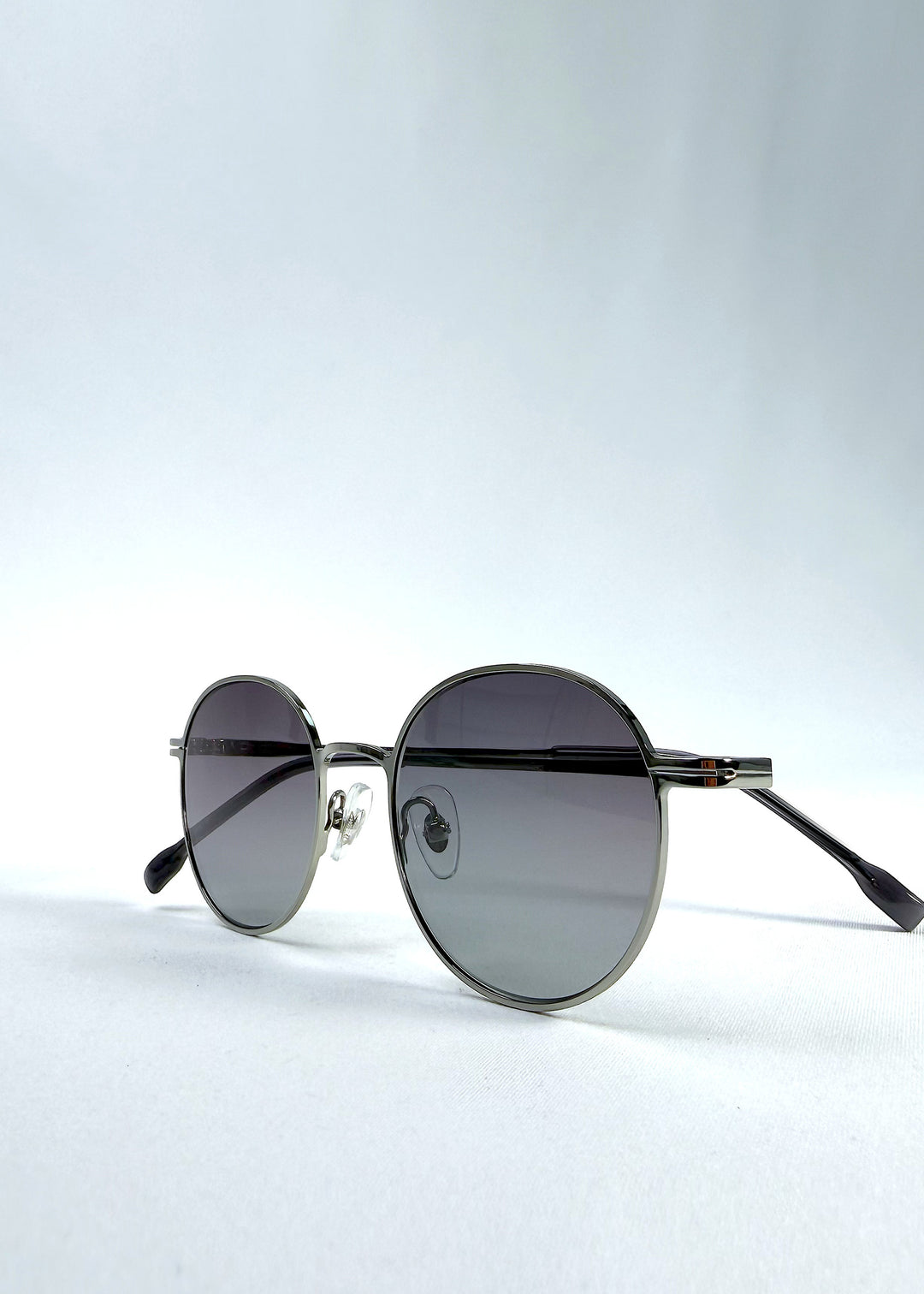 Folded view of AZE Liora Arc sunglasses in Moonlit Silver, featuring a silver-tone round frame and gray gradient lenses, showing the folded temples and frame profile.