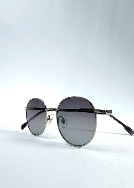 Folded view of AZE Liora Arc sunglasses in Moonlit Silver, featuring a silver-tone round frame and gray gradient lenses, showing the folded temples and frame profile.