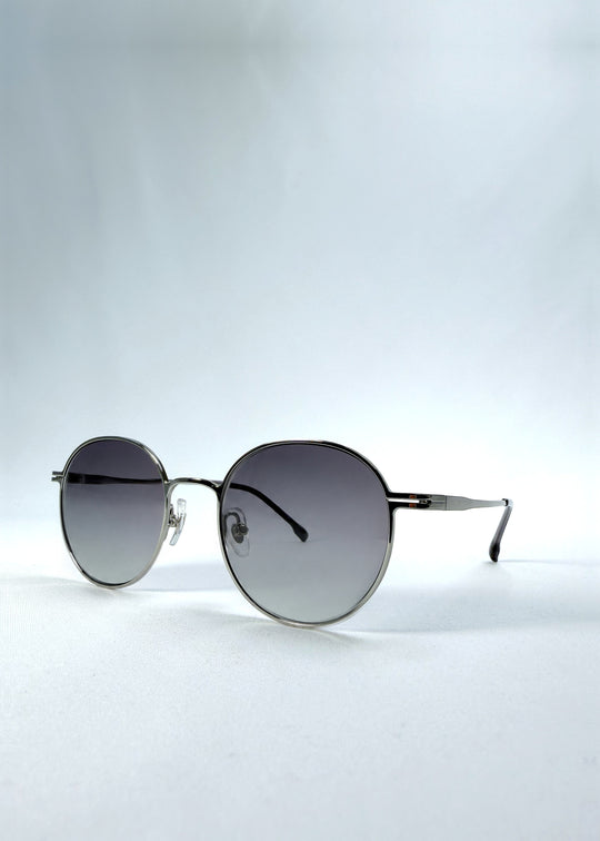 Three-quarter front view of AZE Liora Arc sunglasses in Moonlit Silver, featuring a silver-tone round frame and gray gradient lenses, showing the frame shape and temple profile.