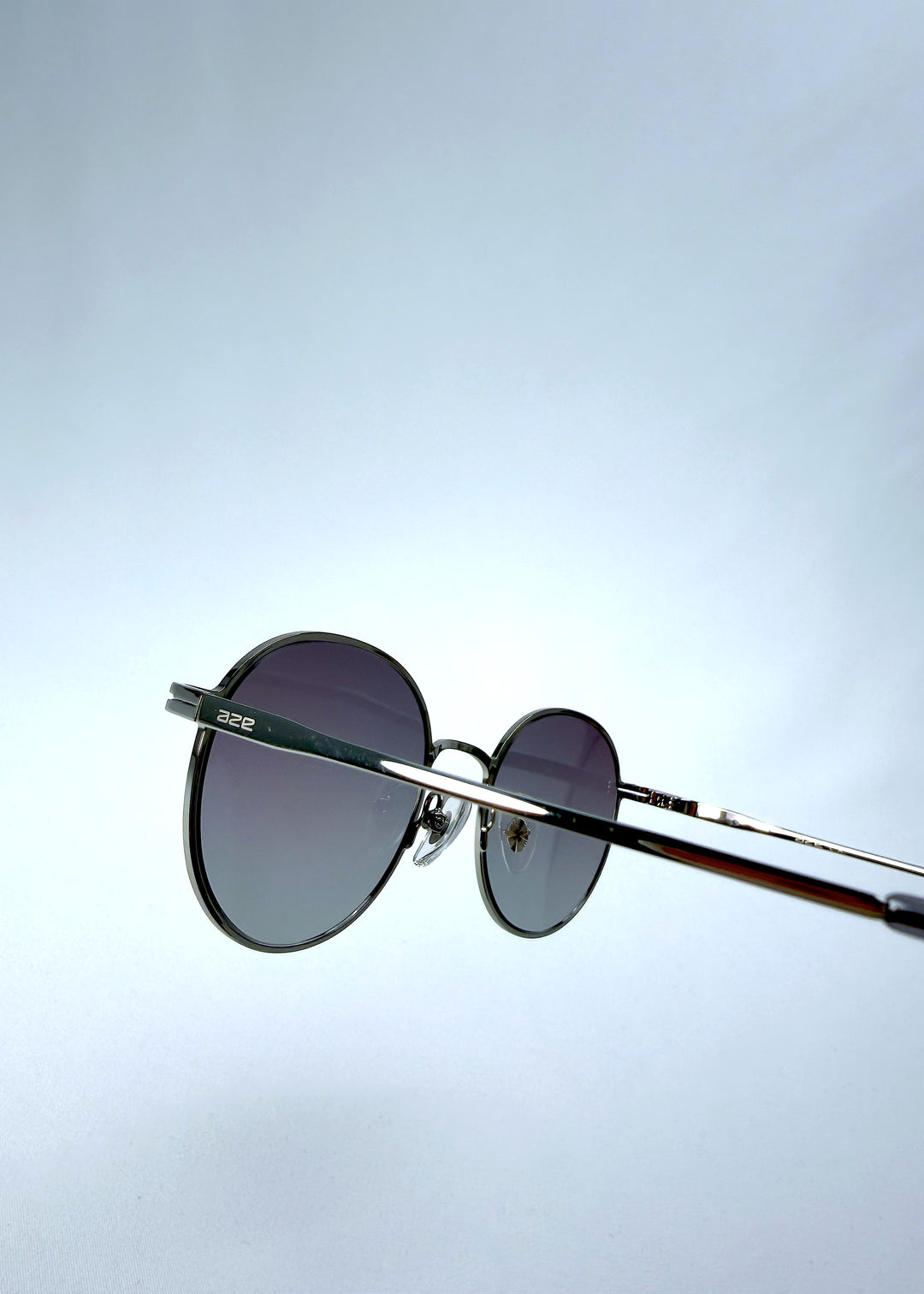 Close-up hinge detail view of AZE Liora Arc sunglasses in Moonlit Silver, featuring a silver-tone round frame and gray gradient lenses, showing the hinge construction, lens edge, and temple connection.