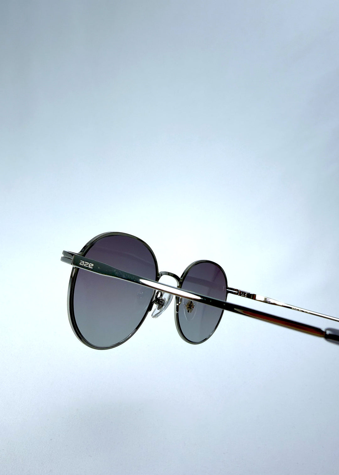 Close-up hinge detail view of AZE Liora Arc sunglasses in Moonlit Silver, featuring a silver-tone round frame and gray gradient lenses, showing the hinge construction, lens edge, and temple connection. 