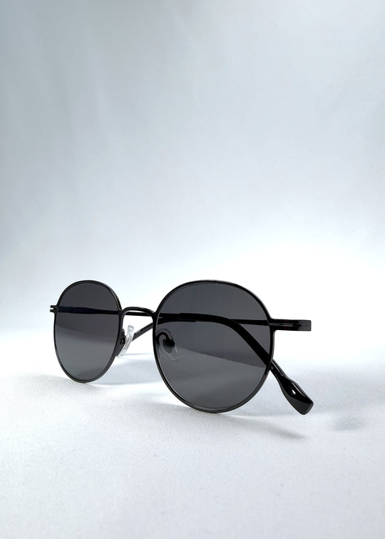 Folded view of AZE Liora Arc sunglasses in Obsidian Black, featuring a slim black round frame and smoky gray gradient lenses, showing the folded temples and frame profile.