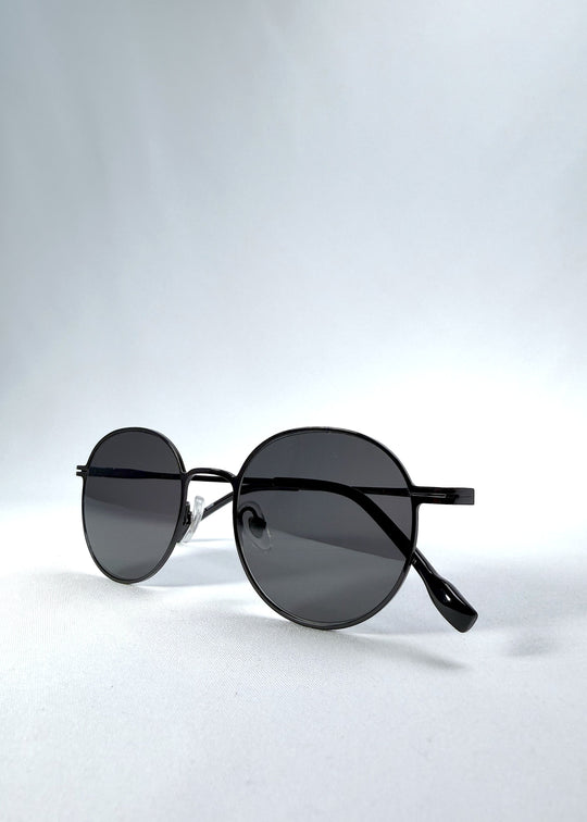 Folded view of AZE Liora Arc sunglasses in Obsidian Black, featuring a slim black round frame and smoky gray gradient lenses, showing the folded temples and frame profile. 