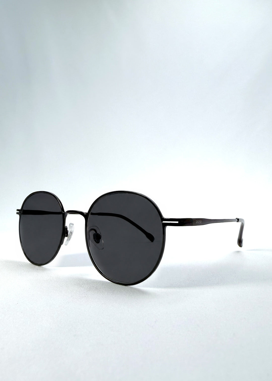 Three-quarter front view of AZE Liora Arc sunglasses in Obsidian Black, featuring a slim black round frame and smoky gray gradient lenses, showing the frame shape and temple profile.