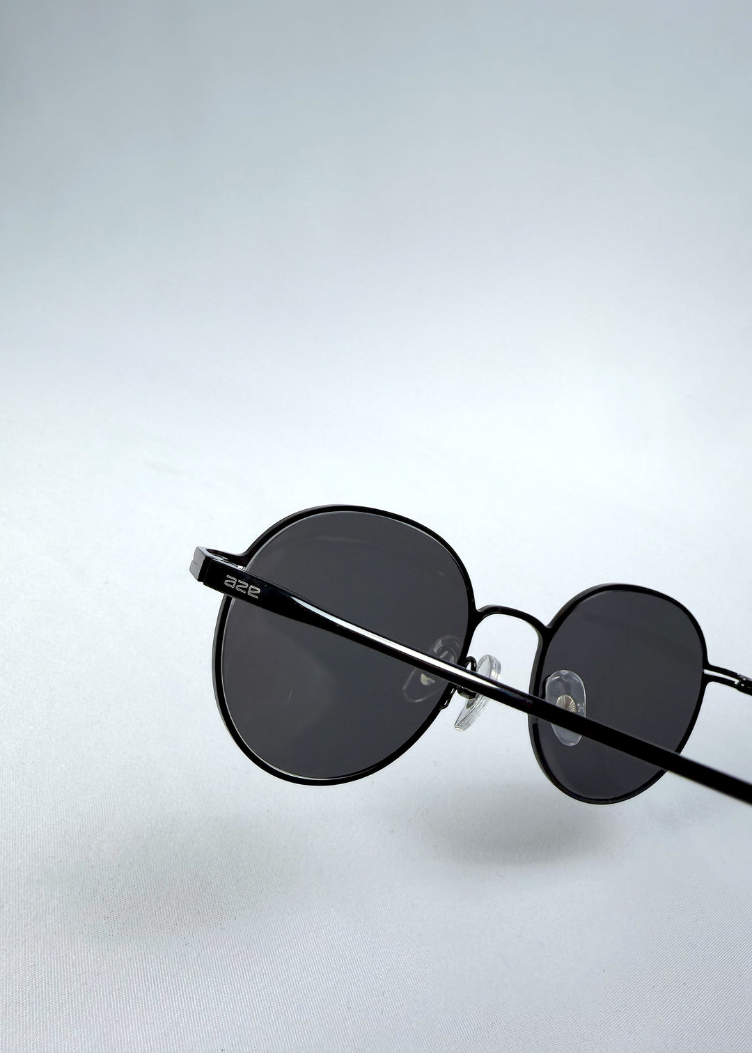 Close-up hinge detail view of AZE Liora Arc sunglasses in Obsidian Black, featuring a slim black round frame and smoky gray gradient lenses, showing the hinge construction, lens edge, and temple connection.