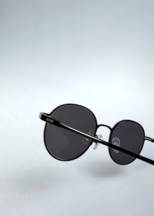 Close-up hinge detail view of AZE Liora Arc sunglasses in Obsidian Black, featuring a slim black round frame and smoky gray gradient lenses, showing the hinge construction, lens edge, and temple connection.