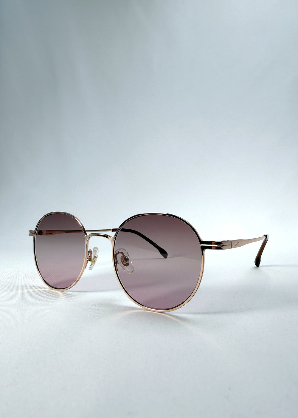 Three-quarter front view of AZE Liora Arc sunglasses in Rosé Dawn, featuring a rose-gold-tone round frame and soft rose gradient lenses, showing the frame shape and temple profile.