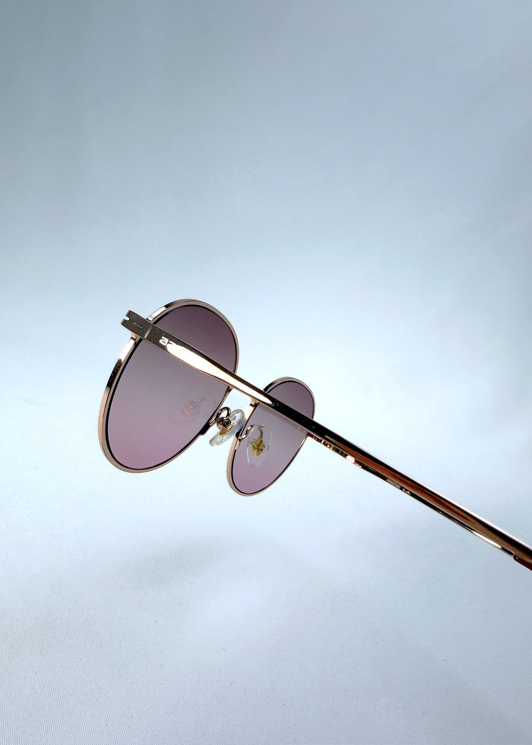 Close-up hinge detail view of AZE Liora Arc sunglasses in Rosé Dawn, featuring a rose-gold-tone round frame and soft rose gradient lenses, showing the hinge construction, lens edge, and temple connection.