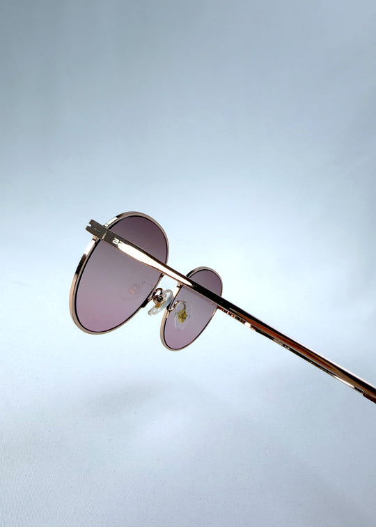 Close-up hinge detail view of AZE Liora Arc sunglasses in Rosé Dawn, featuring a rose-gold-tone round frame and soft rose gradient lenses, showing the hinge construction, lens edge, and temple connection.