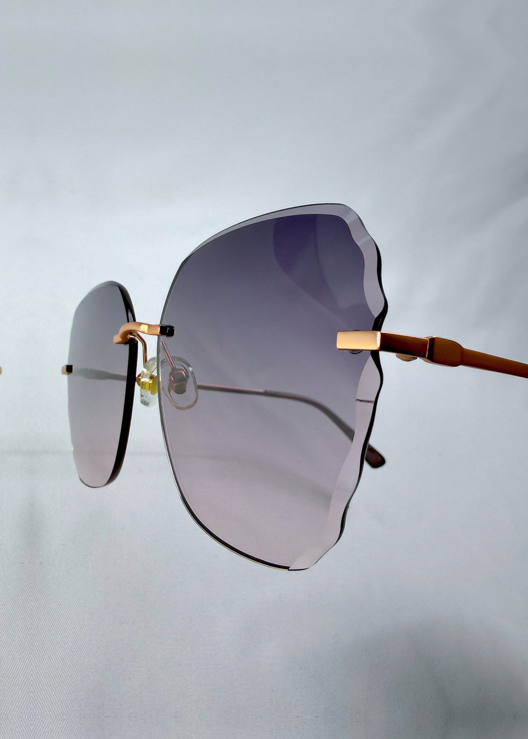 Close-up hinge detail view of Lucera sunglasses in Misty Rosé, featuring rimless gold-tone frame and misty rose gradient lenses, showing the hinge, lens edge, and temple detail. 
