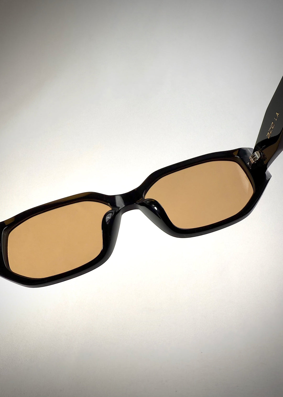 Close-up detail view of Lyra sunglasses in Amber, featuring a glossy black acetate frame with amber-tinted lenses, showing the nose pads and hinge area. 