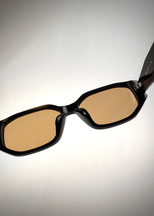Close-up detail view of Lyra sunglasses in Amber, featuring a glossy black acetate frame with amber-tinted lenses, showing the nose pads and hinge area. 