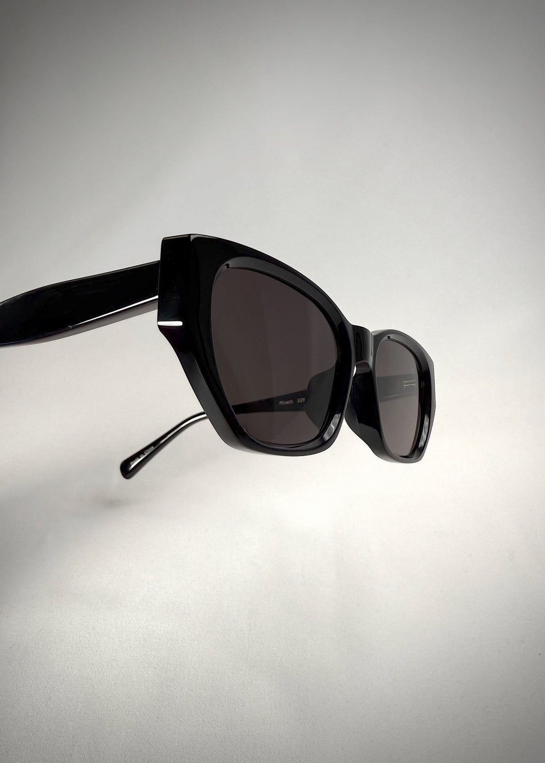 Close-up angled view of Mirach sunglasses in Black, featuring a glossy black acetate frame and gray UV400 lenses, showing the frame edges and glossy finish. 
