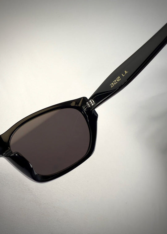 Close-up lens detail view of Mirach sunglasses in Black, featuring a glossy black acetate frame and gray UV400 lenses, showing the lens surface and frame edge. 