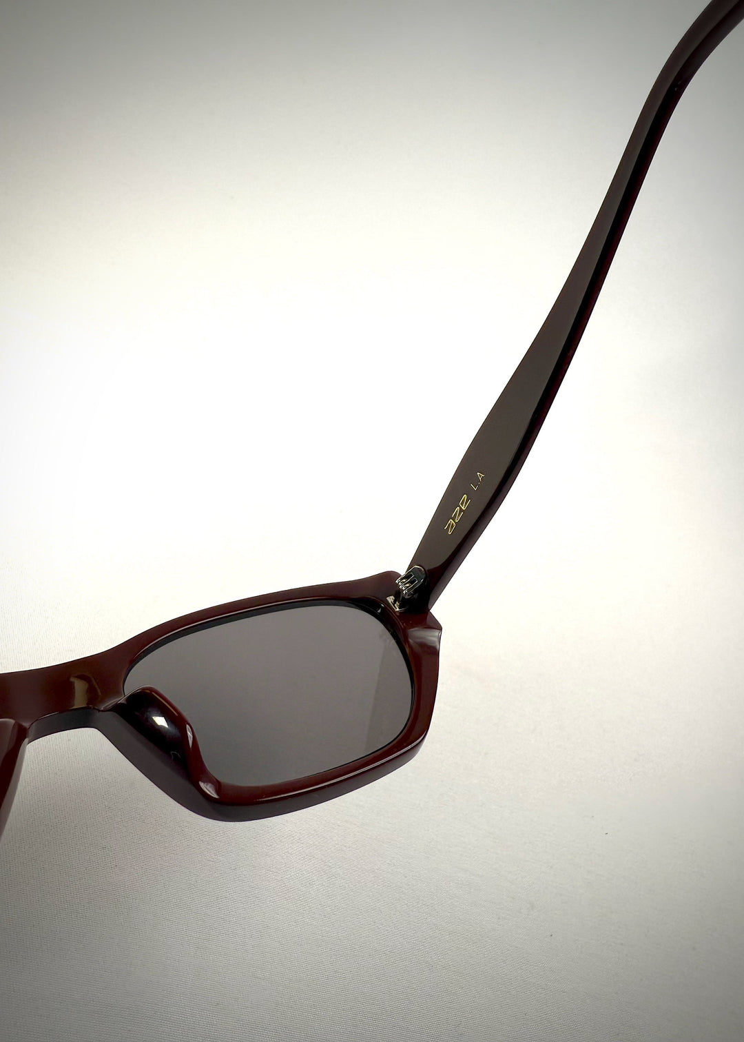 Close-up hinge detail view of Mirach sunglasses in Burgundy, featuring a deep burgundy acetate frame and gray UV400 lenses, showing the hinge construction and lens edge. 
