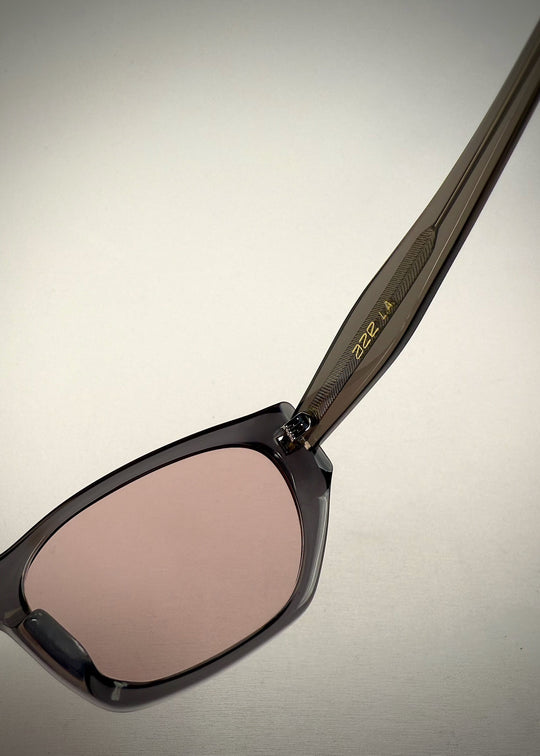 Close-up detail view of Mirach sunglasses in Transparent Gray, featuring a smoke-gray translucent acetate frame and pink-tinted lenses, showing the frame texture and lens edge. 