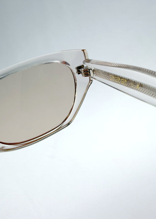 Close-up hinge detail view of Mirach sunglasses in Transparent White, featuring crystal-clear acetate frame and tea-tinted lenses, showing the hinge construction and lens edge.