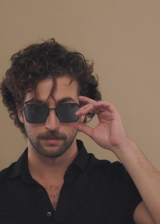 Male model wearing Rigel sunglasses in Blue Graphite with a dark polo, one hand adjusting the temple. 