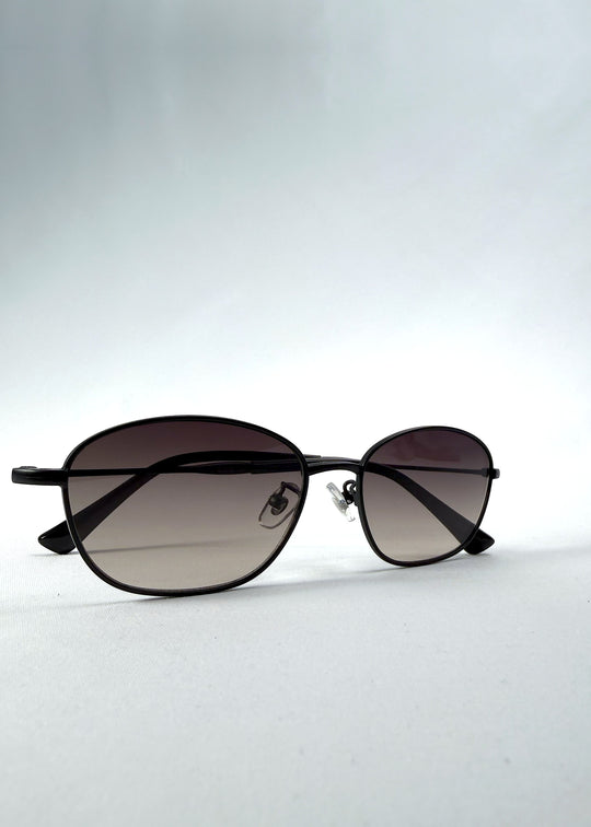 Folded view of Selene Line sunglasses in Matte Black, featuring a slim matte black oval metal frame and smoky gradient lenses, showing the folded temples and frame profile.