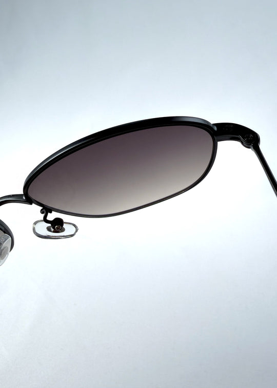 Close-up hinge detail view of Selene Line sunglasses in Matte Black, featuring a slim matte black oval metal frame and smoky gradient lenses, showing the hinge construction, lens edge, and temple connection. 