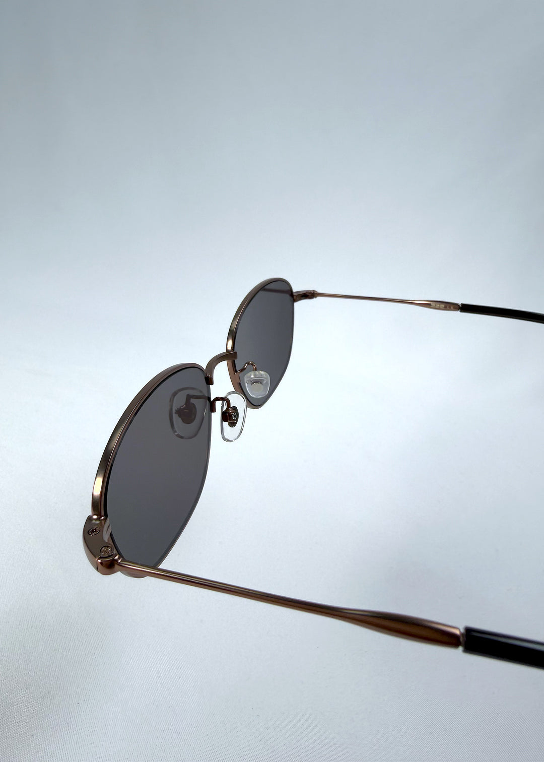 Close-up hinge detail view of AZE Selene Line sunglasses in Mocha Alloy, featuring a slim bronze-tone oval metal frame and smoky brown lenses, showing the hinge construction, lens edge, and temple connection.