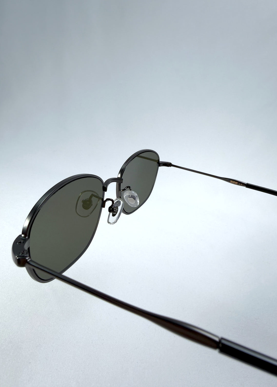 Close-up hinge detail view of AZE Selene Line sunglasses in Sage Gunmetal, featuring a slim gunmetal oval metal frame and sage-green lenses, showing the hinge construction, lens edge, and temple connection.