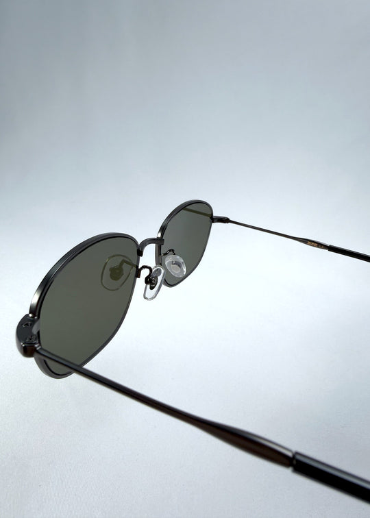 Close-up hinge detail view of AZE Selene Line sunglasses in Sage Gunmetal, featuring a slim gunmetal oval metal frame and sage-green lenses, showing the hinge construction, lens edge, and temple connection. 