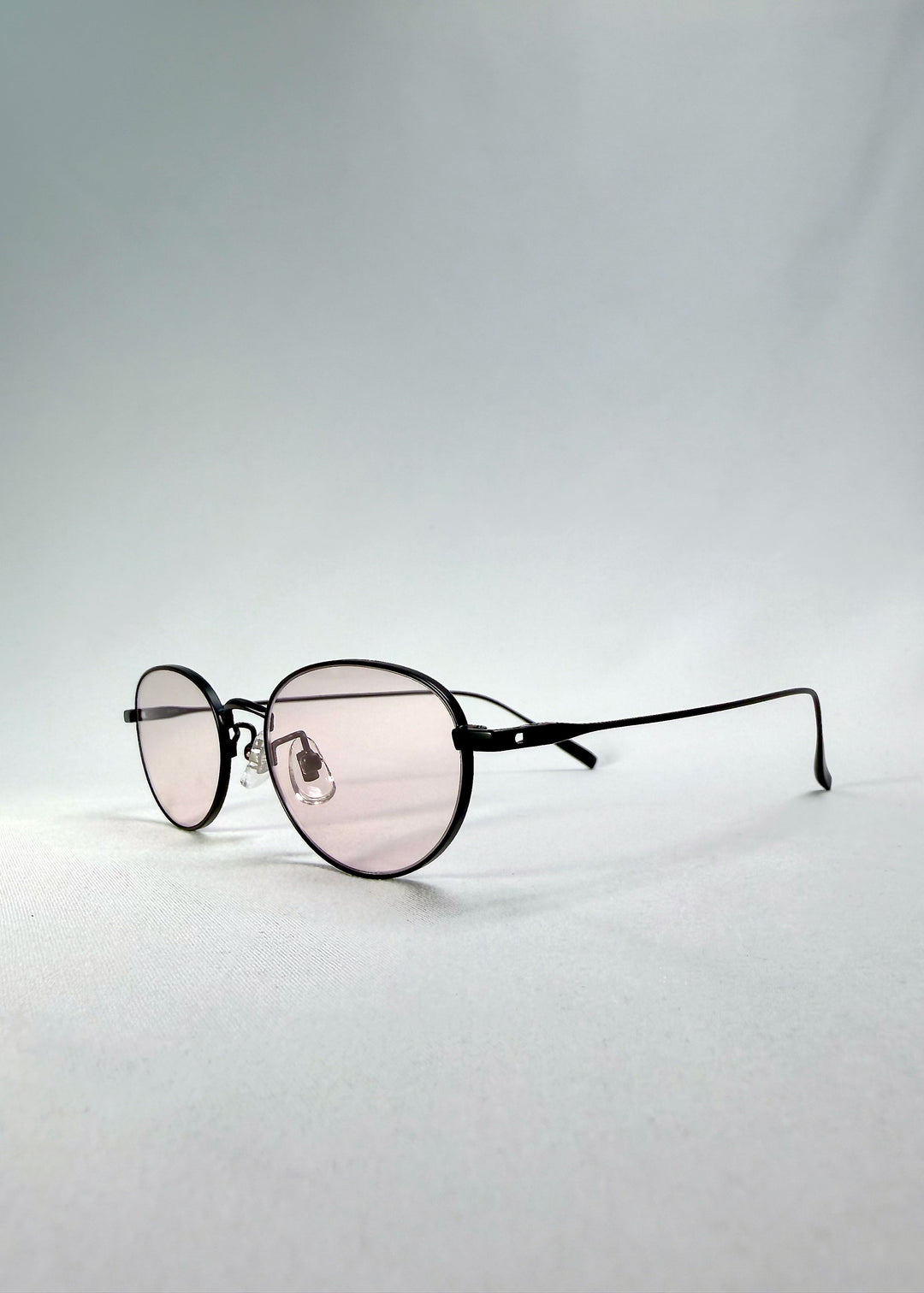 Left front-angle view of Vanta Arc Softint sunglasses in Blush Noir showing the black frame, pale blush lenses, and titanium temples. 