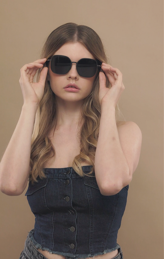 Female model wearing Spica sunglasses in Black with a dark denim top, both hands adjusting the temples. 