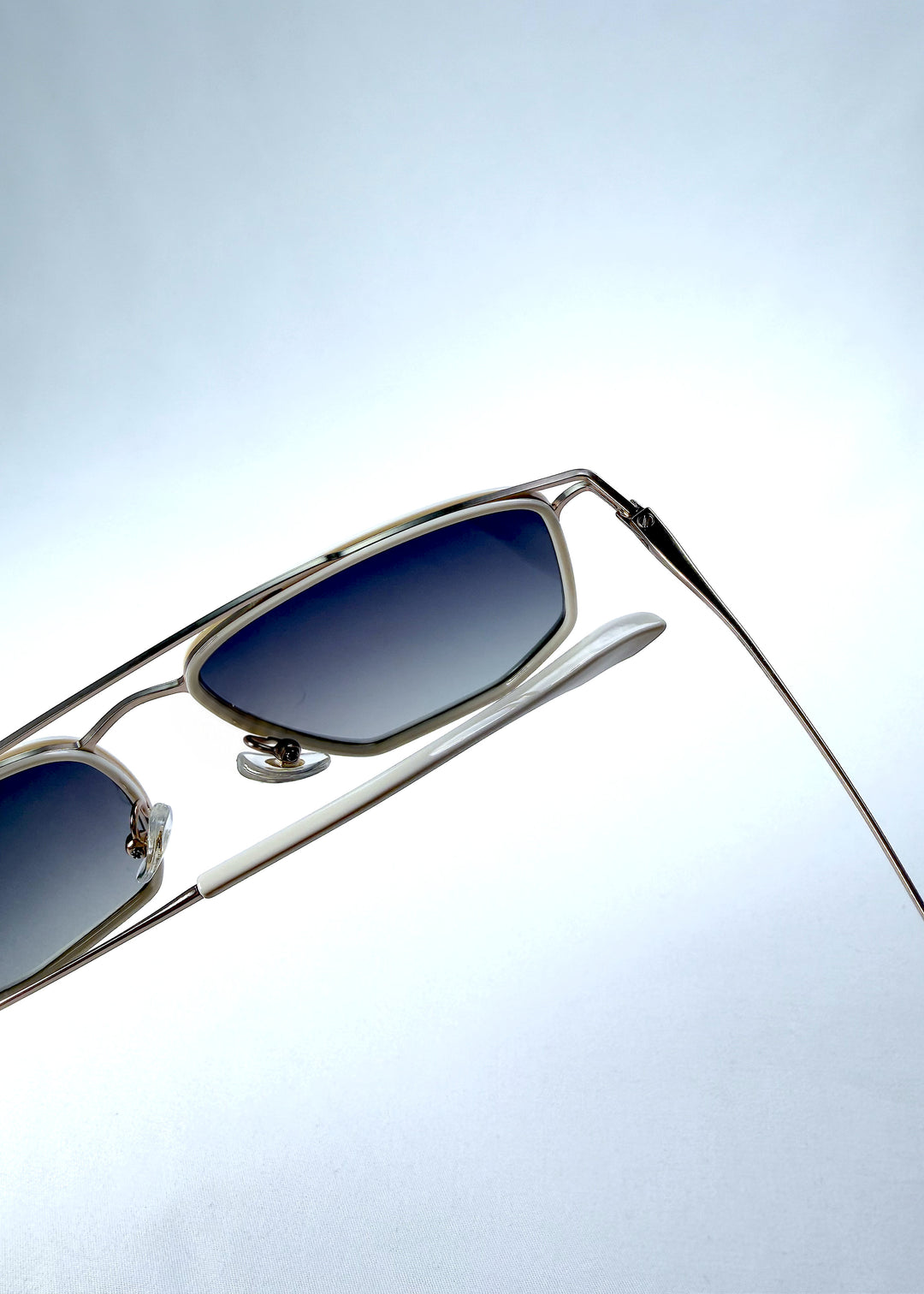 Close-up hinge detail view of AZE Vela Trace sunglasses in Glacial Smoke, featuring a pale gold-tone rectangular frame and blue-gray gradient lenses, showing the hinge construction, lens edge, and temple connection.