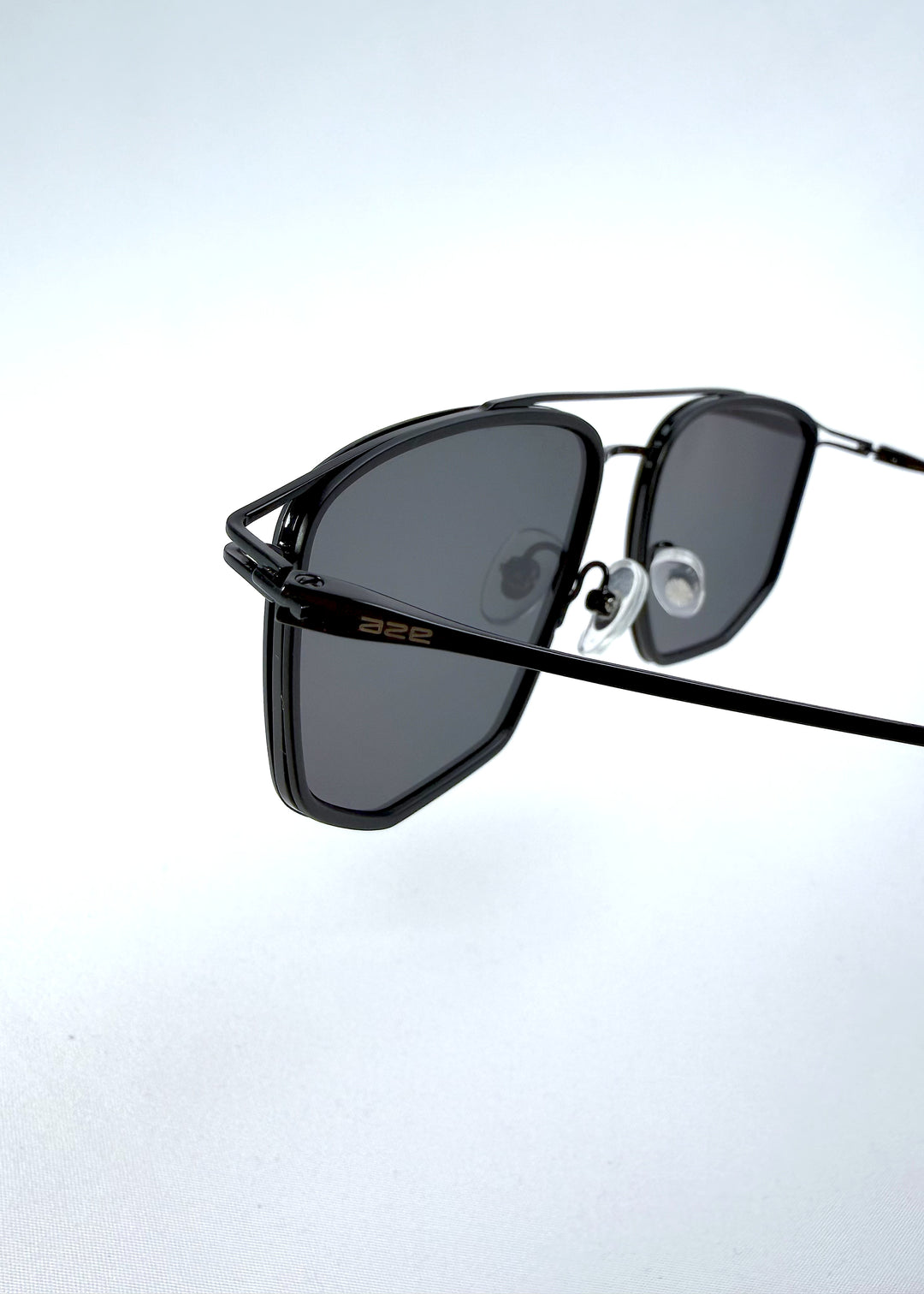 Close-up hinge detail view of AZE Vela Trace sunglasses in Obsidian Black, featuring a structured black rectangular frame and smoky gray lenses, showing the hinge construction, lens edge, and temple connection.