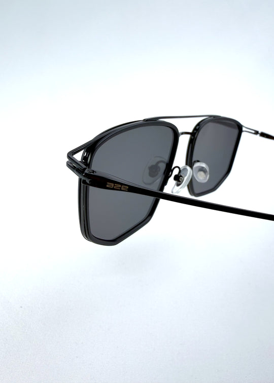 Close-up hinge detail view of AZE Vela Trace sunglasses in Obsidian Black, featuring a structured black rectangular frame and smoky gray lenses, showing the hinge construction, lens edge, and temple connection.