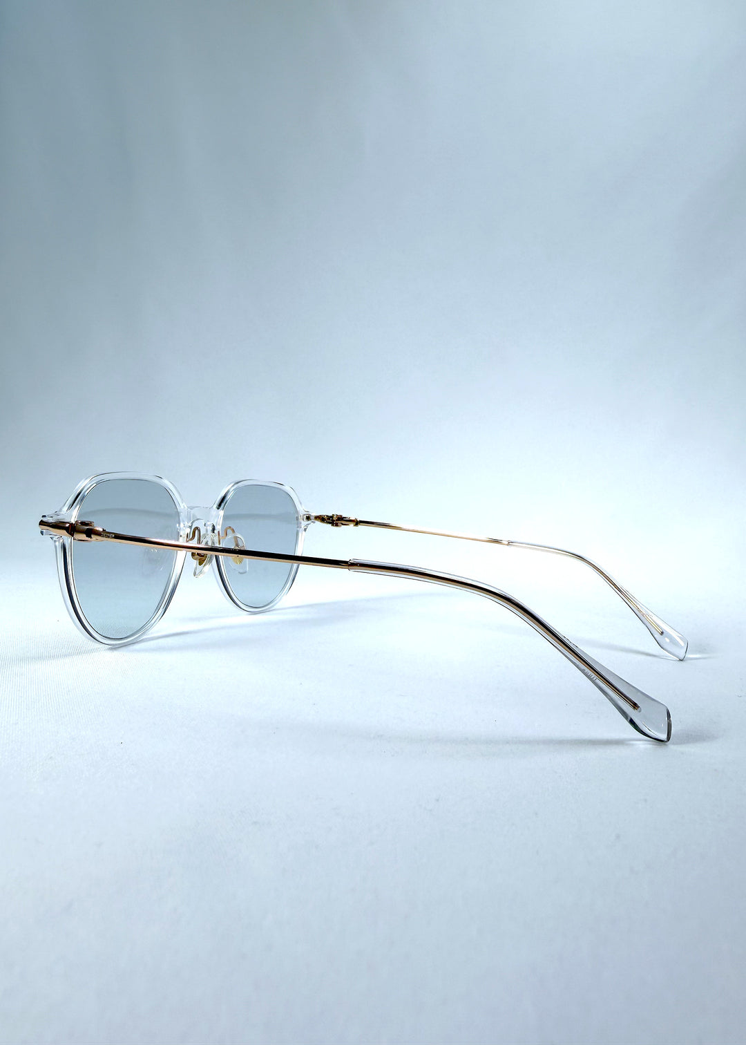 Back-angle view of AZE Vireo Arc sunglasses in Crystal Static, featuring a crystal-clear frame, pale blue lenses, and gold-tone accents, showing the rear frame structure and temple alignment.
