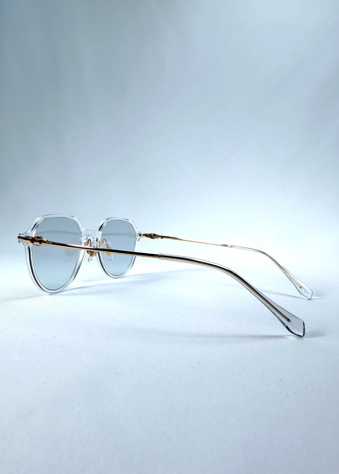 Back-angle view of AZE Vireo Arc sunglasses in Crystal Static, featuring a crystal-clear frame, pale blue lenses, and gold-tone accents, showing the rear frame structure and temple alignment. 
