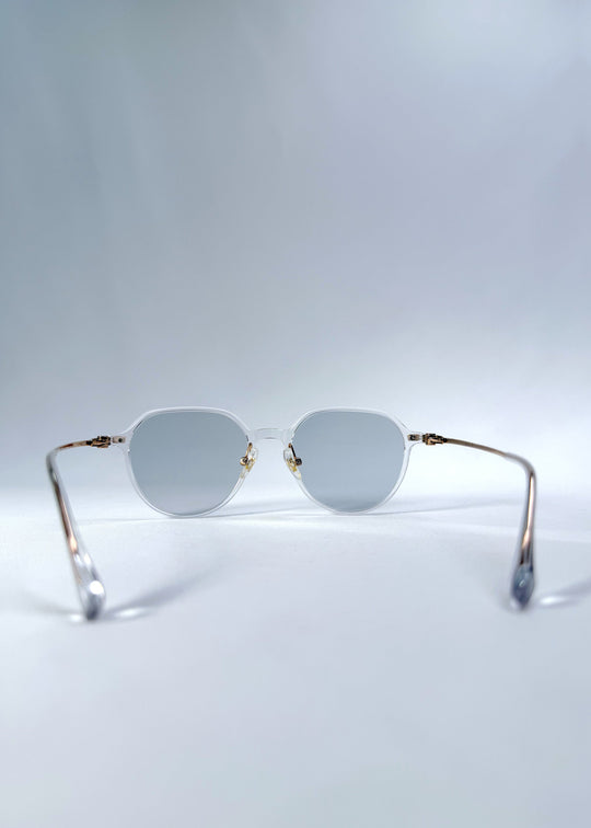 Back view of AZE Vireo Arc sunglasses in Crystal Static, featuring a crystal-clear frame, pale blue lenses, and gold-tone accents, showing the inner frame and open temples. 