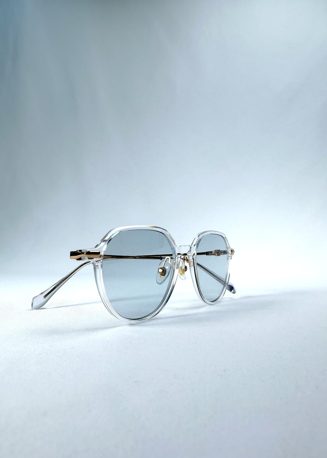 Folded view of AZE Vireo Arc sunglasses in Crystal Static, featuring a crystal-clear frame, pale blue lenses, and gold-tone accents, showing the folded temples and frame profile.