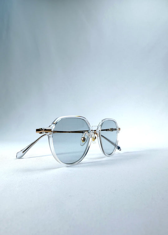 Folded view of AZE Vireo Arc sunglasses in Crystal Static, featuring a crystal-clear frame, pale blue lenses, and gold-tone accents, showing the folded temples and frame profile.